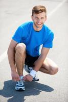 Getting ready to jogging. Top view of man tying shoelaces on sports shoe and smiling while standing outdoors photo