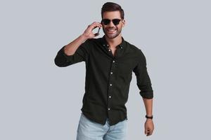 Confident and handsome.  Good looking young man in smart casual wear talking on his smart phone and smiling while standing against grey background photo
