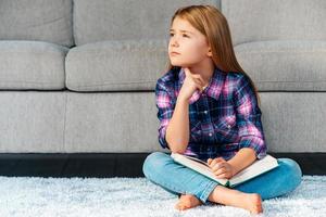 So many questions. Pensive little girl holding book and looking thoughtful while sitting on the carpet in lotus position at home photo