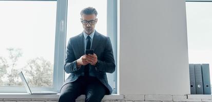 Confident mature man in formalwear using smart phone while sitting on the window sill in office photo