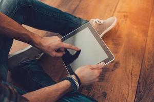 All the world in one touch. Close-up top view of man using digital tablet while sitting on the wooden floor photo
