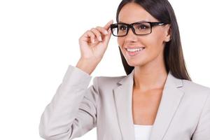 How may I help you Beautiful young businesswoman in suit adjusting her eyeglasses and looking at camera while standing against white background photo