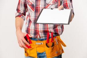 Sign this. Close-up of handyman with tool belt stretching out clipboard with paper standing against grey background photo