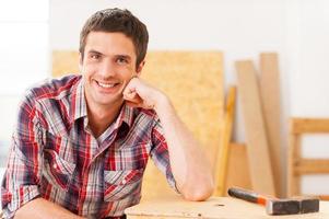 Taking time to relax. Handsome young handyman smiling while sitting in workshop and leaning at the wooden deck photo
