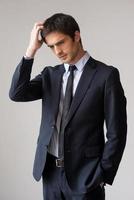 Lost in business thoughts. Thoughtful young man in formalwear holding hand in hair and looking away while standing against grey background photo
