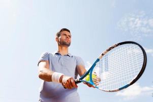 listo para servir. vista en ángulo bajo de un apuesto joven con camisa de polo sosteniendo una raqueta de tenis y una pelota mientras se enfrenta al cielo azul foto