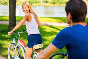 Riding bikes together. Beautiful young smiling woman riding her bicycle and looking over shoulder while her boyfriend riding behind her photo
