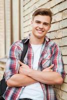 Enjoying time in college. Handsome young man carrying backpack on one shoulder and smiling while leaning at the brick wall photo