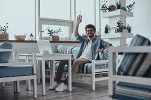 Handsome young modern man looking at camera with smile and waving while sitting on the sofa in restaurant photo