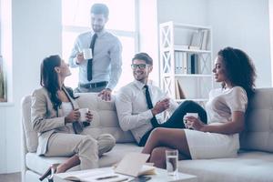 Coffee break. Group of four young people holding coffee cups and discussing something with smile while sitting on the couch at office photo
