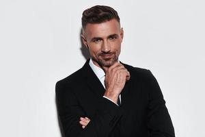 Too good to be real. Handsome young man in full suit keeping hand on chin and looking at camera with smile while standing against white background photo