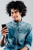 His favorite song. Cheerful young African man wearing headphones and using his smartphone with smile while sitting against grey background photo