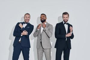 tres apuestos jóvenes en trajes y pajaritas mirando a la cámara y sonriendo mientras están de pie contra el fondo gris foto