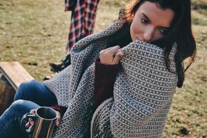 Feeling free and relaxed. Beautiful young woman covered with blanket holding a mug and looking away while spending time by the campfire with her boyfriend photo