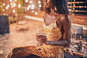 Beautiful young African woman in evening gown holding flute with champagne while spending time on night party photo