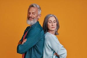 Displeased senior couple keeping arms crossed while standing together against orange background photo