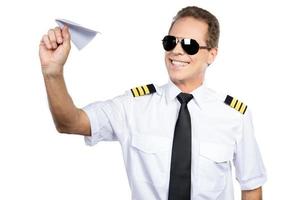 apasionado por su trabajo. piloto masculino confiado en uniforme jugando con un avión de papel mientras está de pie contra el fondo blanco foto