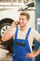 tu coche es como un joven apuesto hablando por teléfono móvil y sonriendo mientras está de pie en el taller con el coche al fondo foto