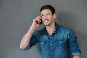 Good talk with friend. Cheerful young man talking on the mobile phone and smiling while standing against grey background photo