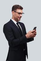 Receiving good feedbacks. Handsome young man using his smart phone and smiling while standing against grey background photo