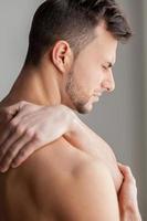 Feeling pain after workout. Rear view of young muscular man touching his shoulder and expressing negativity while standing isolated on grey background photo