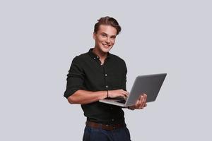 Good looking young man in shirt working on laptop and looking at camera with smile while standing against grey background photo