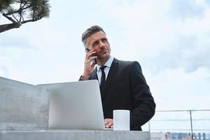 Confident mature man working on laptop and talking on mobile phone while standing on the rooftop terrace photo