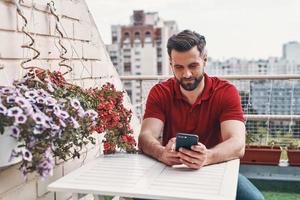 Handsome young man in casual clothing using smart phone while sitting on the rooftop patio photo