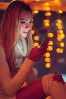 Reading lovely message from him. Beautiful young woman in white sweater holding smart phone and looking at it with smile and with defocused Christmas lights in the background photo