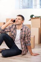 Calling from a new apartment. Handsome young man sitting on the floor and talking on the mobile phone while cardboard boxes laying on the background photo