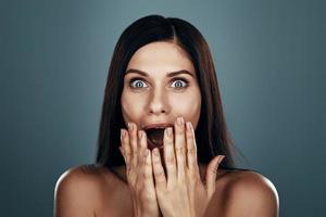 Surprised young woman looking at camera and covering mouth with hand while standing against grey background photo