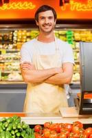 How may I help you Handsome young cashier keeping arms crossed and smiling while standing at supermarket checkout photo
