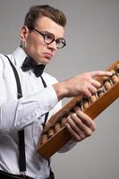 Nerd with abacus. Low angle view of serious young man in bow tie and suspenders using abacus while standing against grey background photo