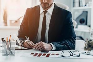 Quick notes. Close-up of young man in full suit writing something in his notebook while sitting at the office desk photo