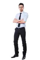 Confident businessman. Full length of handsome young man in shirt and tie keeping arms crossed and looking at camera while standing against white background photo