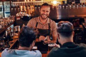 joven barman sirviendo cerveza a hombres jóvenes mientras está de pie en el mostrador del bar en el pub foto