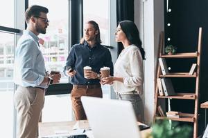 On the break. Young colleagues in smart casual wear discussing something and holding disposable cups while standing in the office photo