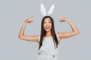 mira a una hermosa joven asiática señalando sus orejas de conejo y sonriendo mientras se enfrenta a un fondo gris foto