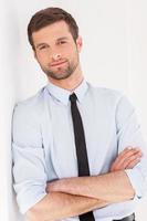 Thoughtful businessman. Handsome young man in shirt and tie looking at camera and keeping arms crossed while leaning at the wall photo