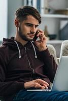 Working from home. Concentrated young man using his laptop and talking on mobile phone while sitting on couch at home photo