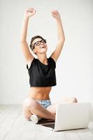 Beautiful winner. Happy young short hair woman keeping arms raised while sitting on the floor with laptop laying near her photo