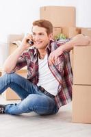 Just moved in. Cheerful young man sitting on the floor and talking on the mobile phone while cardboard boxes laying on the background photo