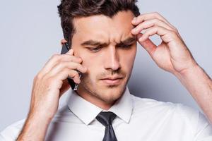 Bad news. Depressed young man in shirt and tie talking on the mobile phone and touching head with hand while standing against grey background photo