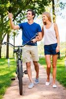 pareja con bicicleta. hermosa joven pareja sonriente caminando con bicicleta en el parque mientras el hombre señala hacia afuera y sonríe foto
