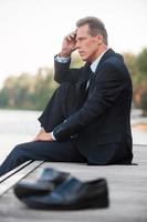 Worried about his business. Side view of thoughtful businessman holding hand on chin and looking away while sitting barefoot at the quayside and with shoes laying on foreground photo