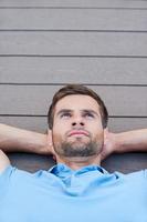 Man daydreaming. Top view of thoughtful young man holding hands behind head and looking away while lying down on the hardwood floor photo