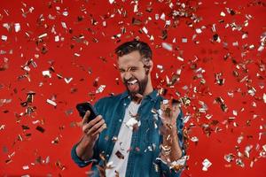 Happy young man in casual clothing holding smart phone and gesturing while standing against red background with confetti flying around