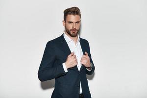 Confident and successful. Handsome young man in full suit adjusting his jacket and looking at camera while standing against grey background photo