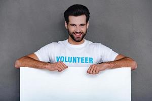 Please donate Confident young man in volunteer t-shirt leaning to white board and looking at camera with smile while standing against grey background photo
