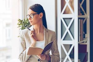 Planning her timetable. Pensive young beautiful businesswoman in glasses holding notebook and looking trough window while standing at her working place photo